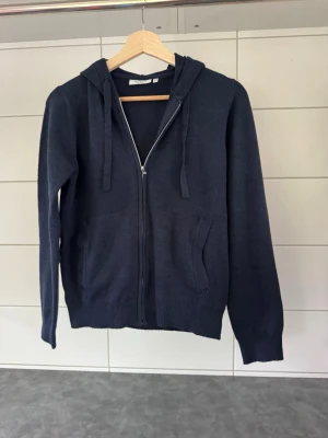 Mörkblå hoodie med dragkedja - Mörkblå hoodie med dragkedja framtill och två fickor. Tröjan har huva med dragsko och ribbade muddar vid ärmslut och nederkant. Perfekt för en avslappnad stil och enkel att matcha med jeans eller joggers.