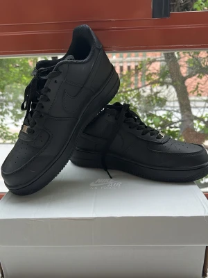 Nike Air Force 1 helsvarta sneakers - Säljer ett par klassiska Nike Air Force 1 sneakers i helsvart läder. Skorna har lågt skaft, perforerad tåbox och ikoniska swoosh-loggan på sidan. Perfekta för en clean och stilren look. Levereras med svarta skosnören och matchande sula.