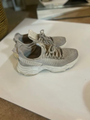 Vita Steve Madden sneakers - Snygga vita sneakers från Steve Madden med stickad ovandel och tjock vit sula. Skorna har snörning och logga på sidan. Perfekta för dig som gillar en sportig och trendig look.
