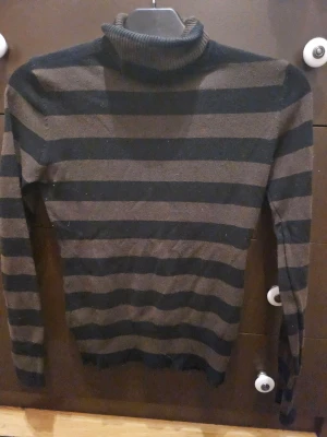 Randig polotröja från H&M - Säljer en stickad polotröja från H&M i svart och mörkbrunt med breda horisontella ränder. Tröjan har lång ärm och ribbad polokrage. Perfekt för dig som gillar en klassisk men ändå chill look.