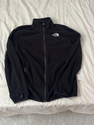 Svart fleecejacka från The North Face - Svart fleecejacka från The North Face med dragkedja framtill och hög krage. Jackan har två sidofickor med dragkedja och diskret logga på bröstet. Perfekt för lager-på-lager och svalare dagar.