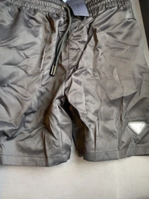 Prada shorts  - Snygga svarta shorts från Prada med klassisk trekantslogga på benet och justerbar snörning i midjan. Tillverkade i ett glansigt syntetmaterial som ger en modern look. Perfekta för dig som vill ha en stilren och exklusiv vibe.