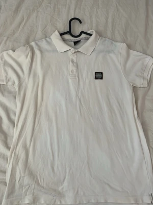 Vit pikétröja från Stone Island - Klassisk vit pikétröja från Stone Island med korta ärmar och krage. Tröjan har två knappar vid halsen och den ikoniska Stone Island-loggan på bröstet. Tillverkad i ett mjukt och bekvämt bomullsmaterial som passar perfekt till jeans eller shorts.
