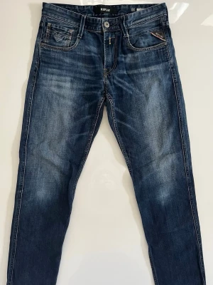 Replay anbass jeans  - Snygga blå slim fit jeans från Replay med klassisk femficksdesign och slitna detaljer framtill och baktill. Jeansen har en mörk tvätt med ljusare partier och kontrastsömmar. Perfekta för en avslappnad och trendig look.