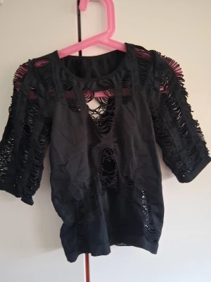 Svart topp med öppen rygg och håldetaljer - Säljer en svart topp med unika håldetaljer och öppen rygg. Toppen har trekvartsärm och är tillverkad i ett tunt material som ger en edgy och trendig look. Perfekt för dig som vill sticka ut och gillar statement-plagg. Storlek XS eller S 