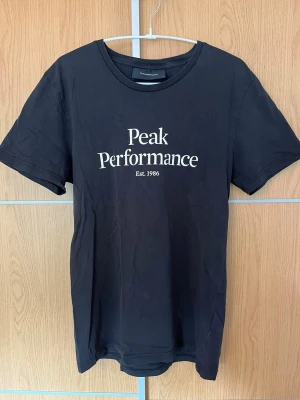 Svart t-shirt från Peak Performance - Svart t-shirt från Peak Performance med vit logotyp och text 'Est. 1986' på bröstet. Klassisk rund halsringning och korta ärmar. Tillverkad i mjuk bomull för en bekväm känsla och enkel stil.