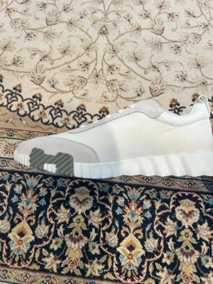 Vita H skor - Säljer ett par stilrena vita H sneakers med ribbad sula och grå logga. Ovandelen är i textil med inslag av mocka vid tån och hälen. Skorna har en modern, låg profil och passar perfekt till streetwear eller casual outfits.