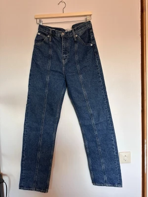 Blå raka jeans från Calvin Klein - Säljer ett par blå raka jeans från Calvin Klein med klassisk femficksdesign och extra fickor bak. Jeansen har hög midja och tydliga sömmar framtill. Materialet är kraftig denim.