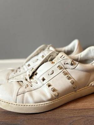 Vita sneakers med nitar från Valentino - Snygga vita sneakers från Valentino med coola silverfärgade nitar längs sidorna och på tungan. Skorna är tillverkade i läder och har klassisk låg profil med vita snören och rund tå. Perfekt för dig som vill sticka ut med en edgy detalj äkta men ingen box därav priset