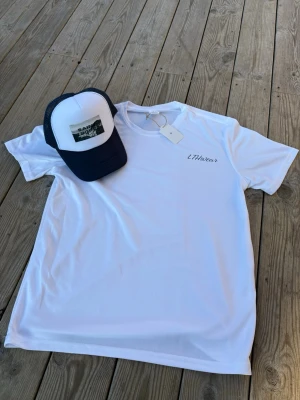 Marin blå keps och Vit t-shirt från LTHwear - Snygg vit t-shirt och marinblå keps från LTHwear med diskret logga på bröstet. Klassisk rund hals och korta ärmar. Materialet känns mjukt och lätt, perfekt för en avslappnad look. Passformen är normal och t-shirten är enkel att matcha med jeans eller shorts.