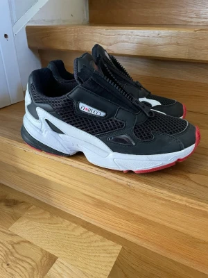 Adidas x Fiorucci svarta sneakers 39 1/3 - Snygga sneakers från Adidas i samarbete med Fiorucci. Skorna är svarta med vita och röda detaljer, mesh- och mockaliknande material samt grov sula. Dragkedja framtill istället för klassiska snören och tydliga loggor på plös och sida. Perfekta för en streetwear-look. Använda sparsamt!