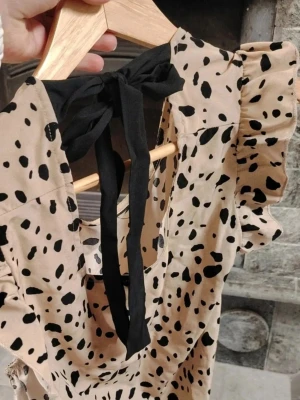 Beige kortklänning med svarta prickar☀️🐆  - Snygg beige kortklänning med svarta prickar och volangärmar😍 Klänningen har en öppen rygg med svart knytband och är tillverkad i material som ger en lyxig men avslappnad känsla. Perfekt för dig som vill sticka ut med en trendig look❤️🐆 Kanske från H&M, men vet ej på säkert. 