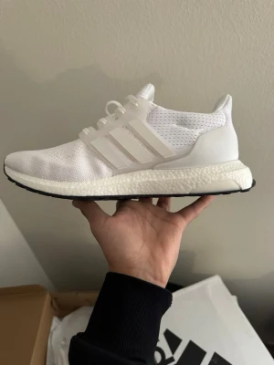 Vita Adidas Ultraboost sneakers - Säljer ett par helvita Adidas Ultraboost sneakers med stickad ovandel i mesh, klassiska tre ränder på sidan och bekväm Boost-sula. Skorna har vita snören och svart yttersula. Perfekta för dig som gillar stilrena och sportiga sneakers.