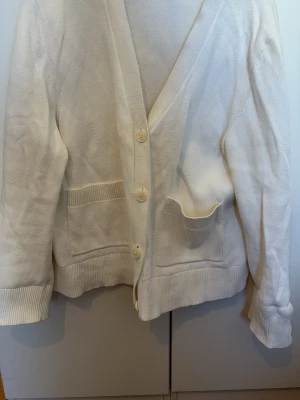 Vit kofta med fickor och knappar - Klassisk vit kofta med tre knappar framtill och två rymliga fickor. Koftan har ribbade muddar vid ärmslut och nederkant, samt en mjuk och bekväm känsla. Perfekt för lager på lager och enkel att matcha med olika outfits.