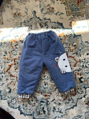 Blå mjukisbyxor med björn-detalj - Mysiga blå mjukisbyxor med elastisk midja och fodrad insida med stjärnmönster. Enat benet har en söt björn-detalj med rutiga öron och rutiga muddar vid bensluten. Perfekta för chill dagar hemma eller när du vill vara extra bekväm.