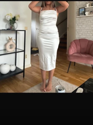Vit satinklänning med smala axelband - Elegant vit satinklänning med smala axelband och figurnära passform. Klänningen har en draperad överdel och slutar strax under knäna. Perfekt för dig som vill ha en stilren och trendig look.