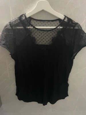 Svart mesh topp med spetsdetaljer - Snygg svart topp med korta ärmar, meshparti upptill och spetsdetaljer. Ovanpå bröstet och axlarna finns transparent mesh med små prickar och spets längs kanterna. 