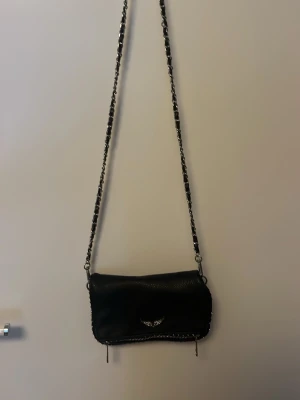 Svart Axelväska från Zadig & Voltaire - Cool svart axelväska i läder från Zadig & Voltaire med silverfärgade detaljer och kedja. Väskan har ett emblem med vingar på framsidan, dragkedja och små nitar längs kanterna. Perfekt för dig som gillar edgy och stilrena accessoarer.