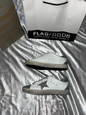 Glittriga Golden Goose sneakers - Säljer ett par vita Golden Goose sneakers med silverglittriga detaljer på häl, tå och stjärna på sidan. Skorna har vita snören med svart text och en robust sula i grått. Perfekta för dig som vill sticka ut med coola och trendiga skor. 24,5