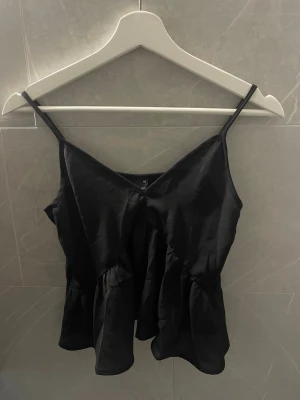 Svart linne  - Svart linne i siden med tunna axelband och v-ringad hals. Toppen har en volangdetalj nedtill som ger ett flowigt och snyggt fall.