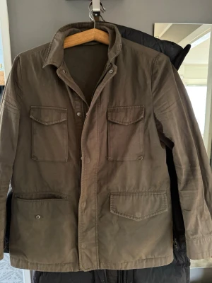 Olivgrön overshirt med fickor - Säljer en olivgrön overshirt  jacka med fyra stora fickor framtill och klassisk krage. Overshirten har knappar och dragkedja samt är tillverkad i ett slitstarkt bomullsmaterial. Perfekt för lager-på-lager och en cool, avslappnad look.