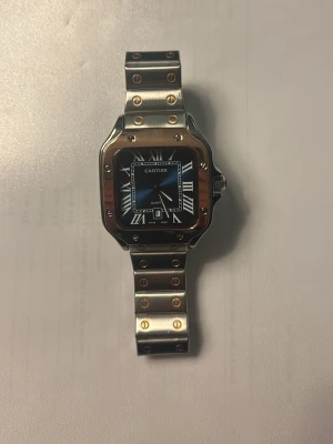  klocka i stål och guld - Snygg Santos klocka med fyrkantig urtavla, romerska siffror och datumvisning. Armband i rostfritt stål med gulddetaljer och skruvar. Klassisk design som sticker ut med sin kombination av silver och guld. Perfekt accessoar för dig som gillar lyxig stil.