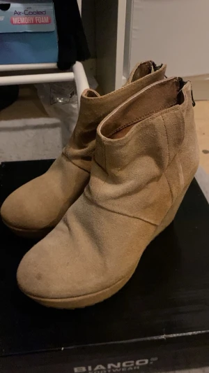 Beige kilklackade stövletter Scorett - Säljer ett par beige stövletter från Scorett med kilklack och dragkedja på sidan. Skorna är tillverkade i mocka och har en stilren, enkel design som passar till många olika outfits. Perfekt för dig som gillar en snygg och bekväm look.