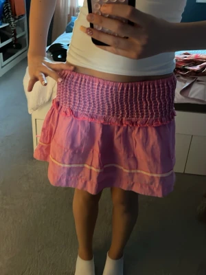 Rosa kortkjol med smock och volang - Säljer en söt rosa kortkjol med bred smock upptill och volangdetaljer. Kjolen är storlek 152 men passar perfekt till XS/S💓💓