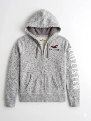 Grå zip-hoodie från Hollister - Snygg gråmelerad hoodie från Hollister med dragkedja, huva och dragsnören. Stor logga på ärmen och broderad logotyp på bröstet. Skick 9/10 inga hål eller defekter