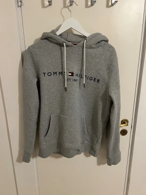 Grå hoodie från Tommy Hilfiger storlek M - Snygg grå hoodie från Tommy Hilfiger med broderad logga och text på bröstet. Klassisk känguruficka framtill, justerbar huva med vita snören och mjukt material. Perfekt för en avslappnad och trendig look.