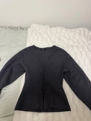 Mörkgrå kofta med knappar från Gina Tricot - Snygg mörkgrå kofta från Gina Tricot med v-ringning och dekorativa knappar framtill. Koftan har lång ärm och är figursydd. Skriv om ni har frågor💗