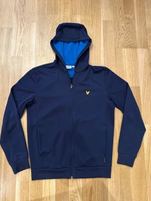 Mörkblå hoodie från Lyle & Scott - Snygg mörkblå hoodie med dragkedja från Lyle & Scott. Den har en blå kontrastfärgad insida på huvan och det klassiska gula fågel-logotypen på bröstet. Materialet är mjukt och stretchigt, perfekt för chill eller träning. Långa ärmar och ribbade muddar.