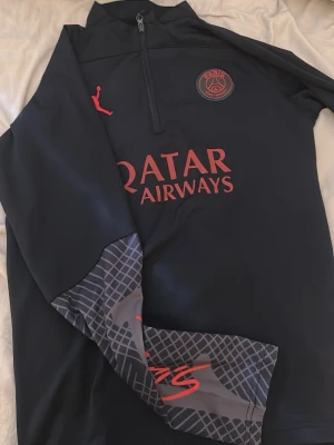 PSG x Jordan svart träningströja - Helt ny!Mörkblå långärmad träningströja från PSG x Jordan med halv dragkedja, röda loggor och Qatar Airways-tryck på bröstet. Ena ärmen har grått mönster och röd signaturdetalj. Tillverkad i ett stretchigt funktionsmaterial, perfekt för fotbollsträning.