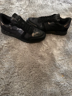 Valentino Garavani Rockrunner Sneakers - Snygga sneakers från Valentino Garavani i svart och grått camomönster. Skorna har lågt skaft, svarta skosnören och detaljer i mocka och läder. Dem är använda men i bra skick, det finns vissa deffekter därav bra pris. Perfekta för dig som vill ha en exklusiv och trendig look.