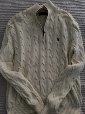 Vit kabelstickad tröja Polo Ralph Lauren - Kabelstickad vit tröja från Polo Ralph Lauren med halv dragkedja och broderad logga på bröstet. Tröjan har hög krage och ribbade muddar. Perfekt för lager-på-lager och klassisk stil.