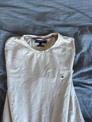 Vit T-shirt från Tommy Hilfiger - Klassisk vit T-shirt från Tommy Hilfiger med rund halsringning och liten broderad logga på bröstet. T-shirten har korta ärmar och är gjord i mjuk bomull, perfekt för en clean och stilren look.