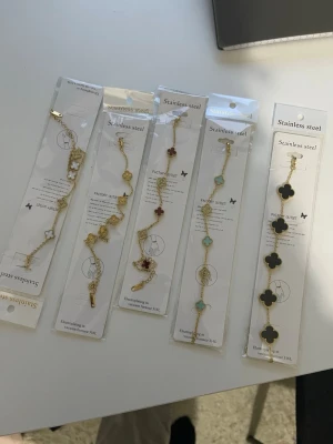 Armband i rostfritt stål, guldpläterade 18+5cm - Säljer flera armband i rostfritt stål med fyrklöverformade pärlor i olika färger som vitt, guld, rött, turkos och svart. Alla armband har guldfärgade länkar och detaljer. Perfekta för att ge din outfit en trendig touch.