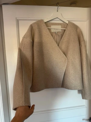 Beige ullkappa från AWARE - Stilren beige kappa från AWARE med rak, boxig passform och markerade sömmar. Kappan har v-ringad hals, är ofodrad och tillverkad i mjuk ullmix. Perfekt för dig som gillar minimalistisk och modern stil.
