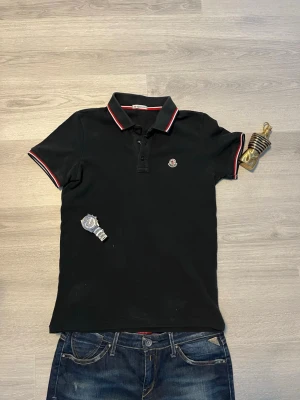 Svart Moncler pikétröja med röd/vit kant - Snygg svart pikétröja från Moncler med klassisk krage och knappar framtill. Tröjan har röda och vita detaljer på krage och ärmslut samt Moncler-logga broderad på bröstet. Perfekt för en clean och stilren look. Funkar som S men är storlek xs