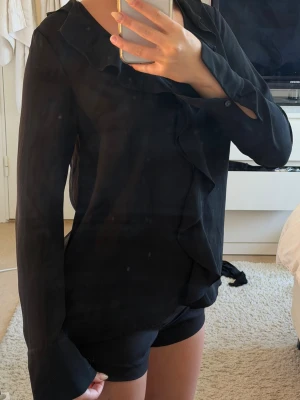 Svart blus med volangdetaljer - Stilren svart blus med långa ärmar och snygga volangdetaljer längs v-ringningen och framtill. Blusen har en lös passform och är tillverkad i ett tunt, mjukt tyg som ger ett elegant fall. Perfekt att styla med både jeans och kjol.
