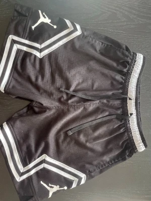 Svarta Jordan shorts med vita detaljer - Snygga svarta Jordan shorts med vita och grå ränder samt klassisk Jumpman-logga på båda benen. Shortsen har snörning i midjan och är tillverkade i ett ventilerande, perforerat polyestermaterial. Perfekta för sport eller chill. Storlek: Det står M men passar bättre på S 📏 Frakt: Snabb och billig frakt 🚚 💴 
