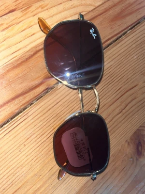Ray-Ban solglasögon med guldiga bågar - Snygga Ray-Ban solglasögon med unika, fyrkantiga linser i mörkbrunt och tunna guldiga metallbågar. Klassisk Ray-Ban-logga på linsen och bekväma skalmar. Perfekta för att lyfta din stil under soliga dagar. Kom priv för pris och vid snabb affär kan jag sänka 