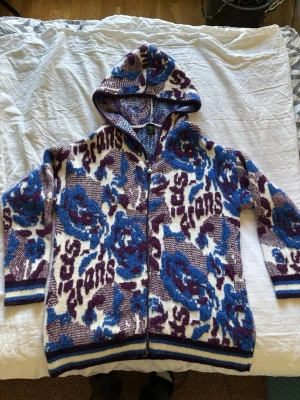 Mönstrad hoodie med dragkedja - Unik hoodie med dragkedja och huva, stickad i tjockt material. Färgerna är lila, blå och vit med ett stort blommönster och textdetaljer. Ribbstickade muddar i nederkant och ärmslut med blå och vita ränder. Perfekt för dig som gillar statement-plagg och streetwear.