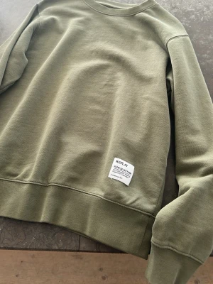 Olivgrön sweatshirt från Replay - Snygg olivgrön sweatshirt från Replay med rund halsringning och ribbade muddar vid ärmslut och nederkant. Tröjan har en vit patch med Replay-logga på framsidan och är tillverkad i mjukt bomullsmaterial. Perfekt för en avslappnad och stilren look.