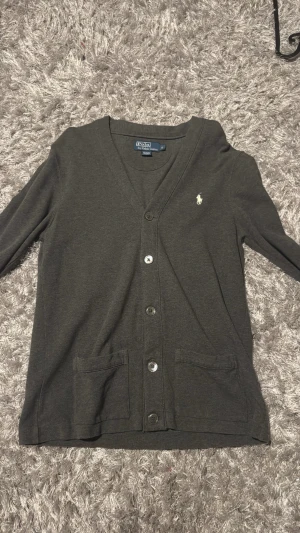 Mörkgrå kofta från Polo Ralph Lauren - Mörkgrå kofta med v-ringning från Polo Ralph Lauren. Klassisk design med knappar framtill, två fickor och broderad logotyp på bröstet. Materialet är mjukt och tunt, perfekt för lager-på-lager. Enkel och stilren look.