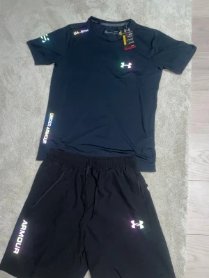 Under armour set - Säljer ett svart träningsset från Under Armour med t-shirt och shorts. Båda plaggen har logotypen och text i regnbågsfärgad stil. Materialet är lätt och snabbtorkande, perfekt för sport och gym. T-shirten har rund hals och korta ärmar, shortsen har elastisk midja och dragsko. Helt ny.