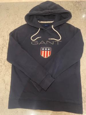 Mörkblå hoodie från GANT med tryck - Mörkblå hoodie från GANT med vit dragsko och broderad logga samt sköld med stjärnor och ränder på bröstet. Klassisk passform och mjukt material, perfekt för en avslappnad stil. Hoodien har långa ärmar och ribbade muddar.