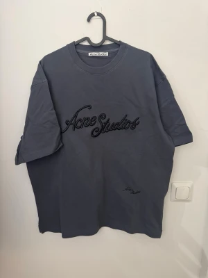 Grå Acne Studios t-shirt med broderi - Snygg grå t-shirt från Acne Studios med stort broderat logotyp på bröstet och mindre broderi nedtill. Oversized passform och klassisk rund halsringning. Perfekt för dig som gillar stilrena plagg med en twist.
