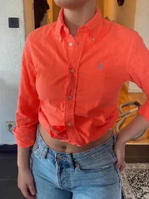 Korallfärgad skjorta från Ralph Lauren - Säljer en snygg korallfärgad skjorta från Ralph Lauren med klassisk button-down krage och blå logga på bröstet. Skjortan är i bomull, har långa ärmar och knappar framtill. Perfekt för dig som vill ha en färgklick i garderoben! Ett litet hål på axeln längst upp som syns knappt😁 Köpt över 2000kr