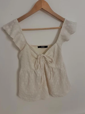 blus med snörning från Bik Bok - Säljer en söt off white blus från Bik Bok med volangärmar och snörning framtill. Blusen har ett subtilt blommigt mönster i tyget och är tillverkad i ett lätt bomullsmaterial. Passar xs-s